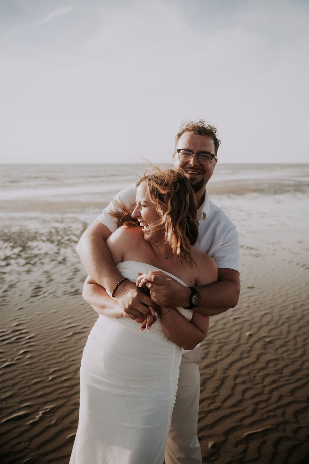 A couple embraces on a sandy beach, the woman in a white dress and man in white shirt, sharing a joyful moment by the ocean.
