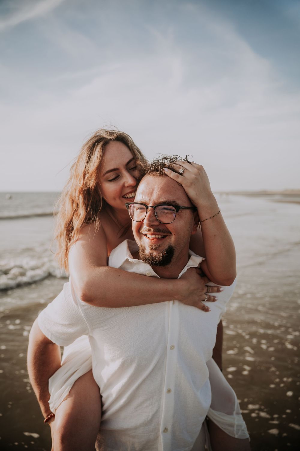 A couple shares a joyful piggyback ride on the beach, with the woman kissing the smiling man's head.