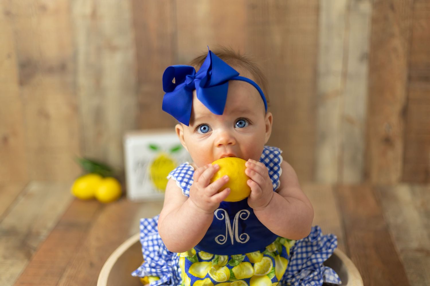 Baby in blue dress and bow holding yellow fruit against wooden background.