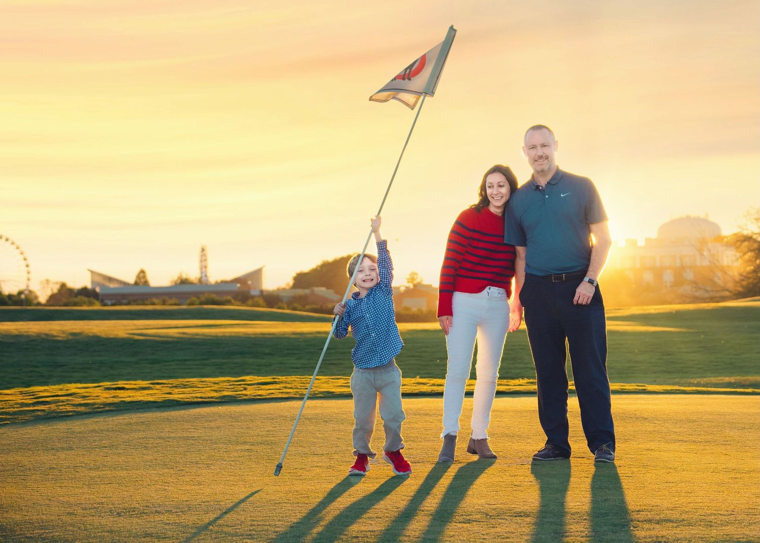Three people stand together on a golf course at sunset with long shadows cast on the green beside a flagstick.