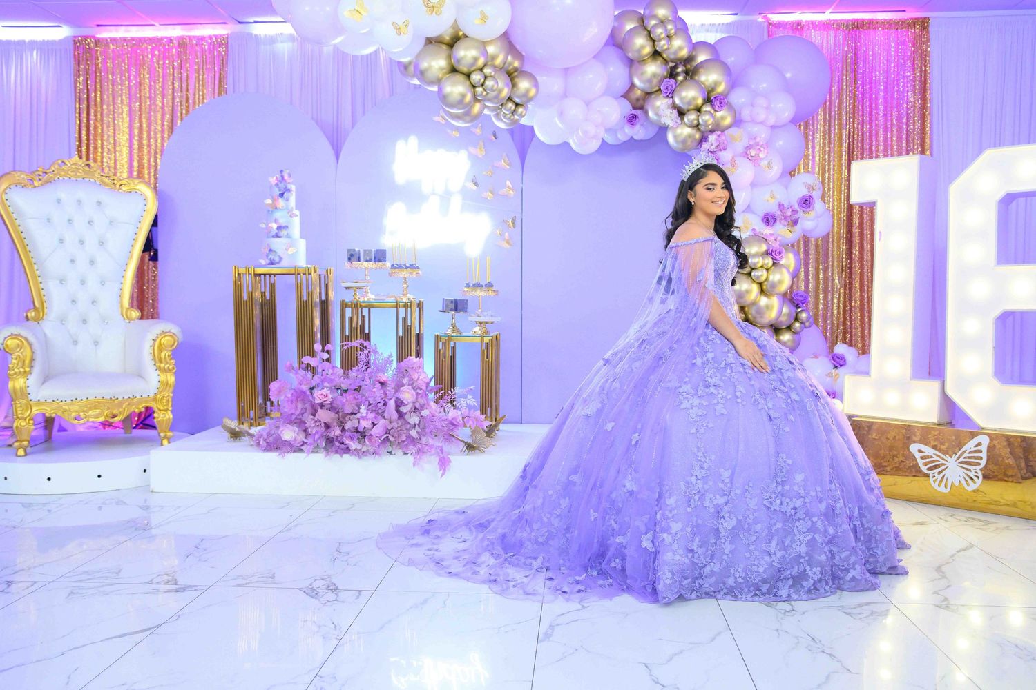 Lavish quinceañera celebration featuring purple decor with floral arrangements, white throne chair and elegant ballgown in matching theme.