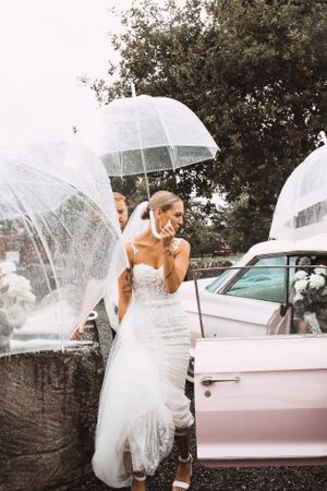 Bride with clear umbrella next to pink vintage car in rainy weather.