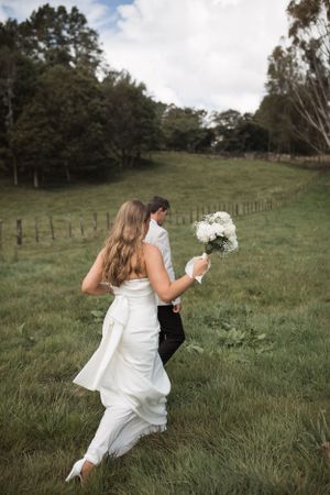 Couple running through a grassy field with white bouquet on their wedding day.