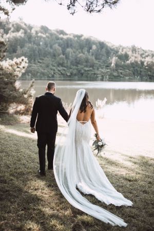 A couple walking hand in hand by a serene lake with a flowing white wedding dress trailing behind.