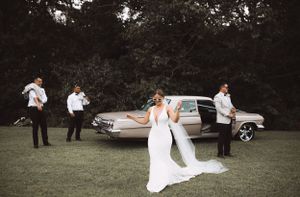 Wedding party posing casually around a classic car on a grassy lawn.