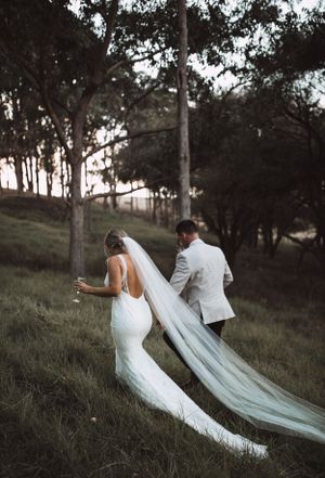 Couple in wedding attire walking up a hill with flowing veil trailing behind.