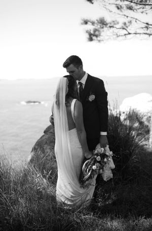 A romantic black and white wedding photo of a couple embracing on a cliff overlooking the ocean.