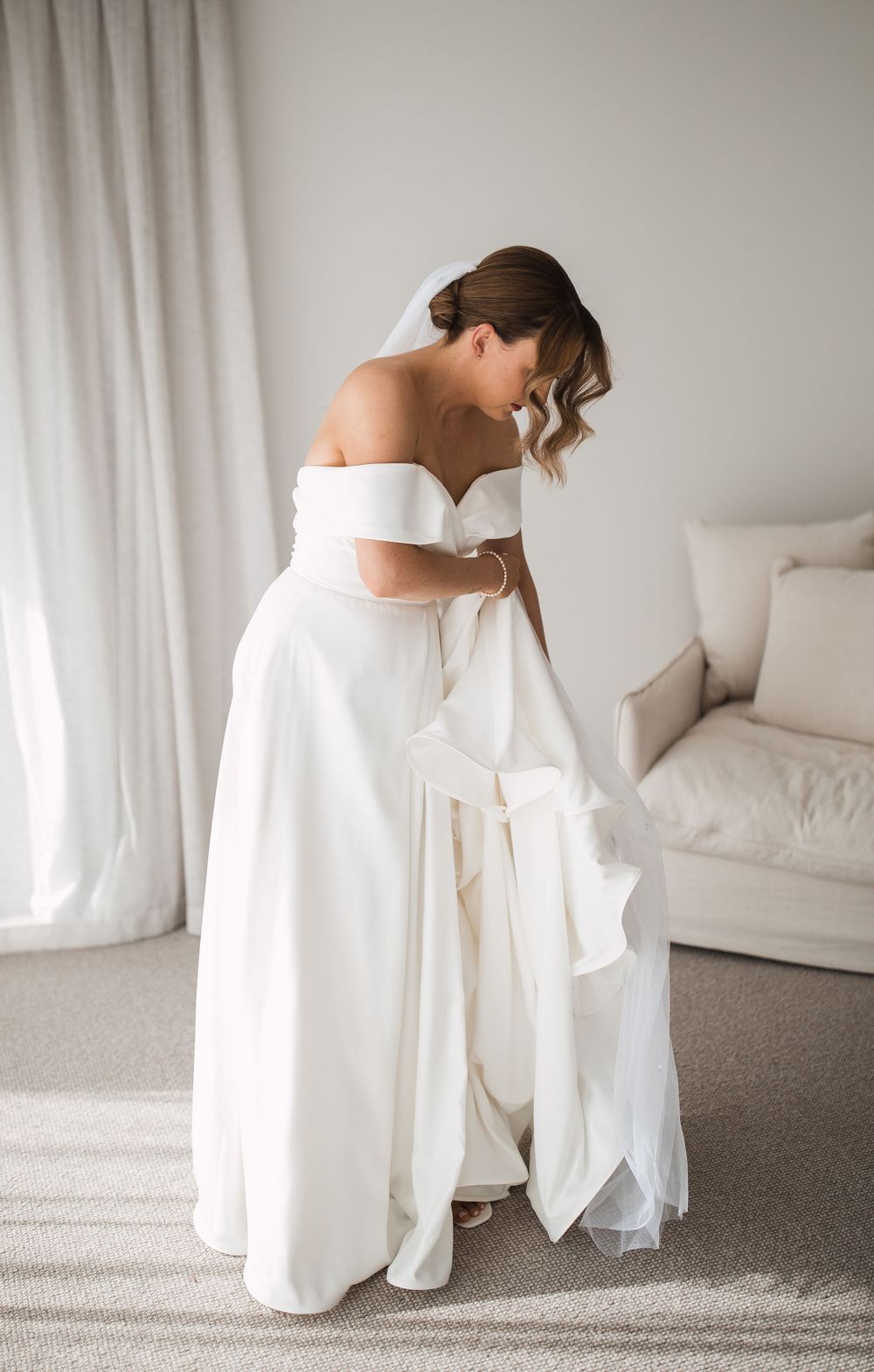 Bride in elegant off-shoulder white wedding gown preparing in a light-filled room.