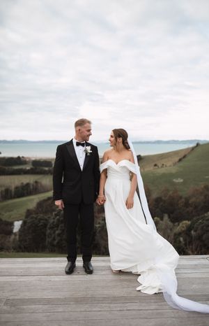 Couple in wedding attire standing on wooden deck with scenic rolling hills in background.