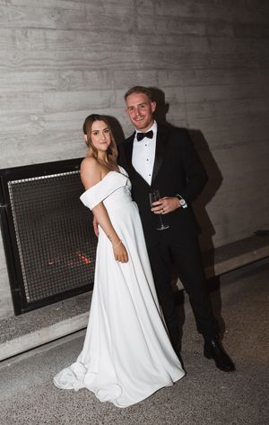 Elegant couple in formal wedding attire posing by modern fireplace at reception.