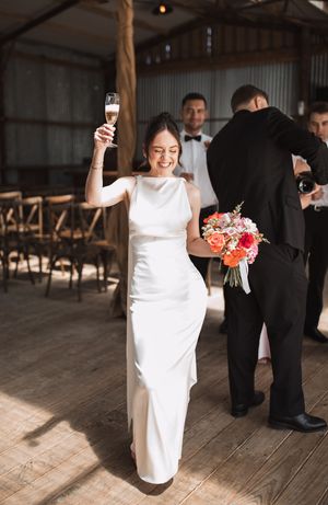 Bride in sleek white dress celebrating with champagne at rustic indoor reception.