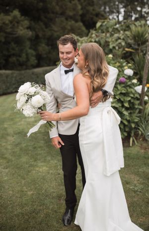 Couple embraces joyfully in garden setting with white floral bouquet.