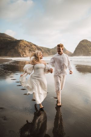 Couple in white outfits runs playfully along a scenic beach with mountains in the background.