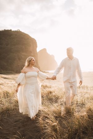 Wedding couple walking through tall grass in a field at sunset.