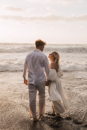 Couple walking along a beach shore during sunset in wedding attire.