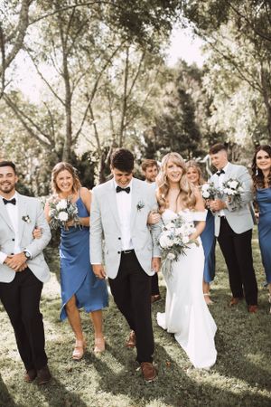Wedding party walks together outdoors wearing blue dresses and white tuxedo jackets.