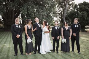 Wedding party in black and white attire poses on a green lawn with trees in the background.