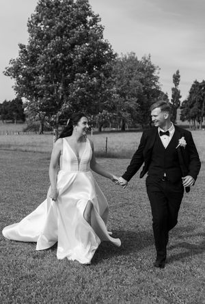 A couple in wedding attire walks playfully through a grassy field on their special day in black and white photography.