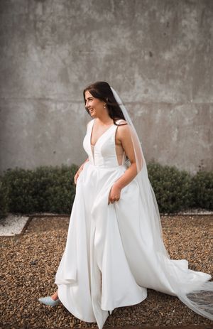 A bride in a flowing white wedding dress spins joyfully on a gravel surface against a concrete wall backdrop.