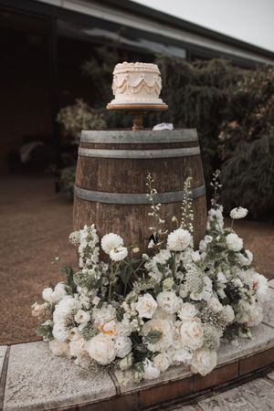 A rustic wooden barrel decorated with white flowers and topped with an elegant wedding cake creates a romantic outdoor display.