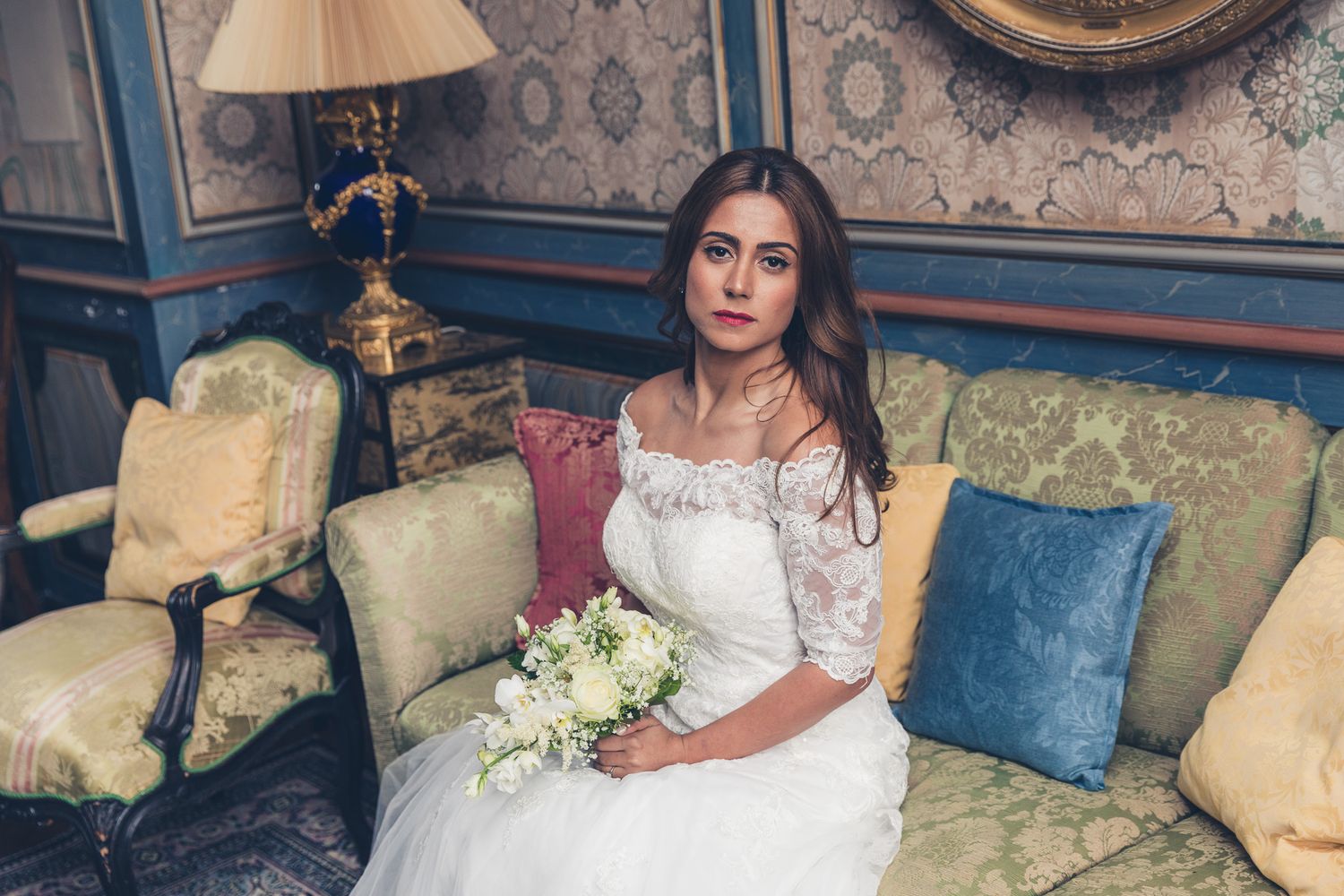 Elegant bride in lace dress holding white bouquet while seated on vintage sofa.