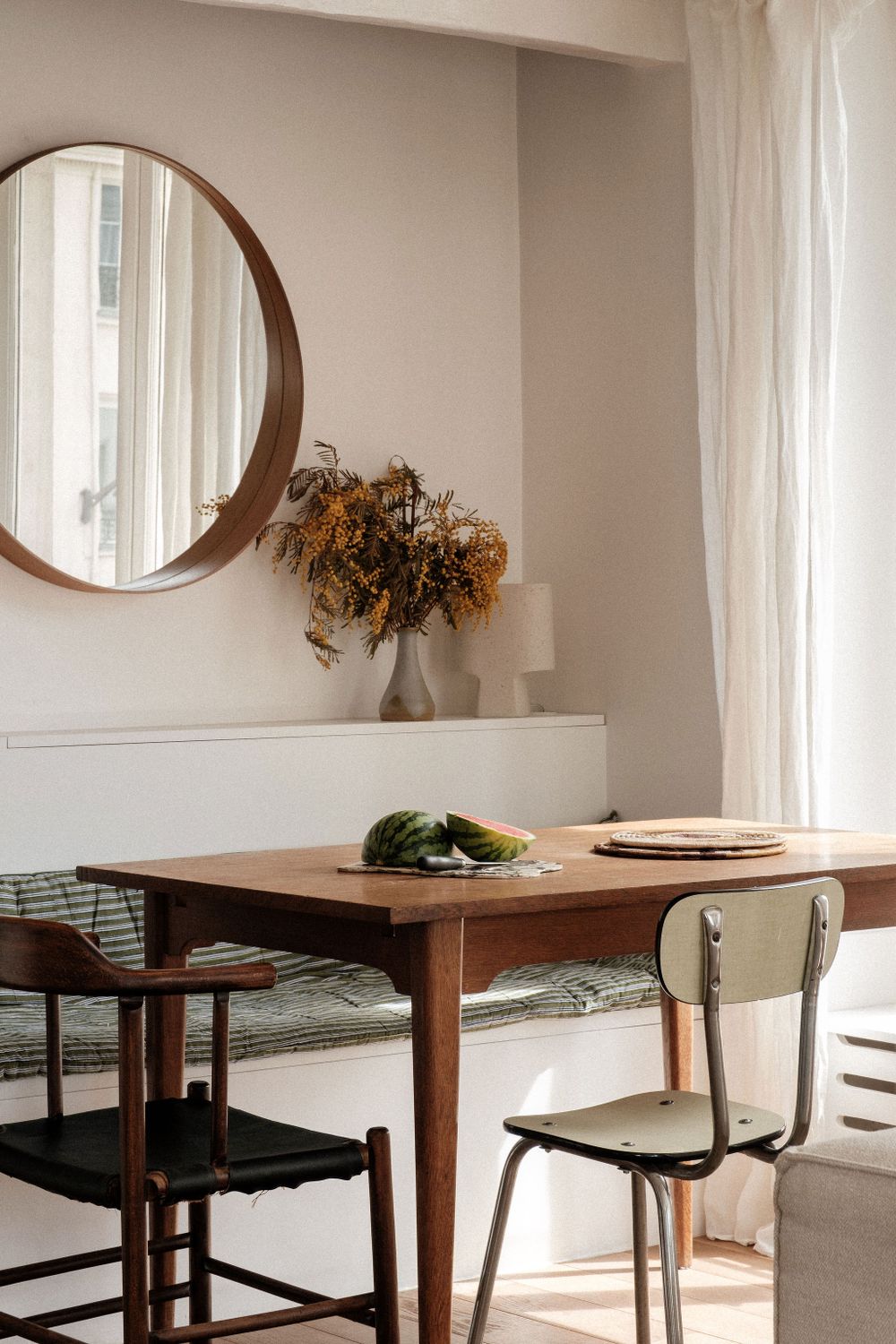 Dining area with round mirror, wooden table, and dried flowers in natural light.