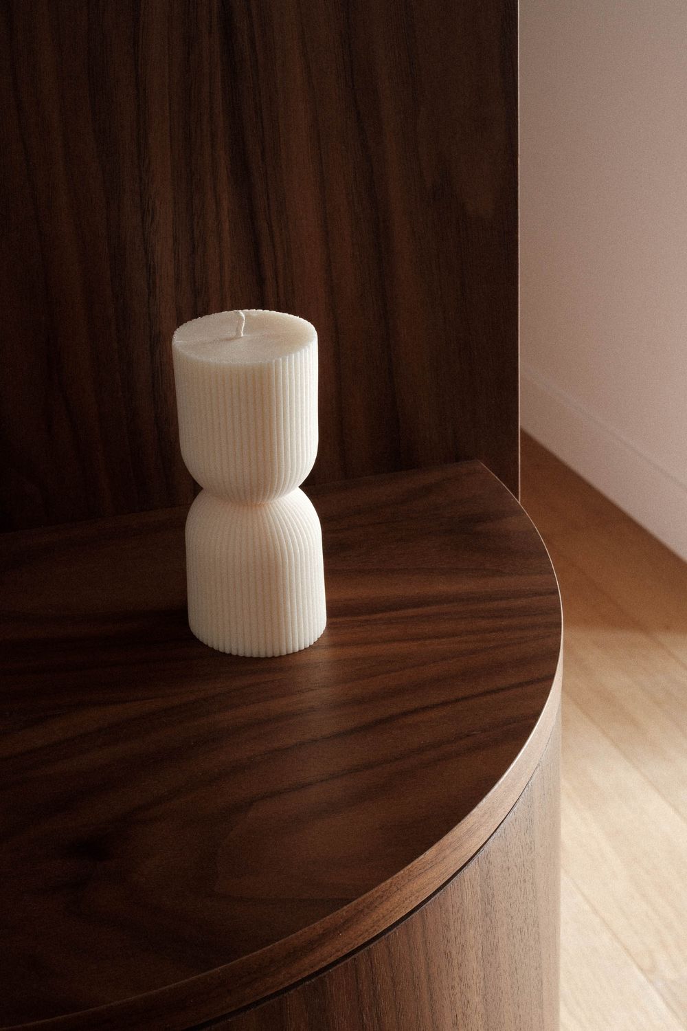 White ribbed candle sitting on dark wooden curved surface in minimalist setting.