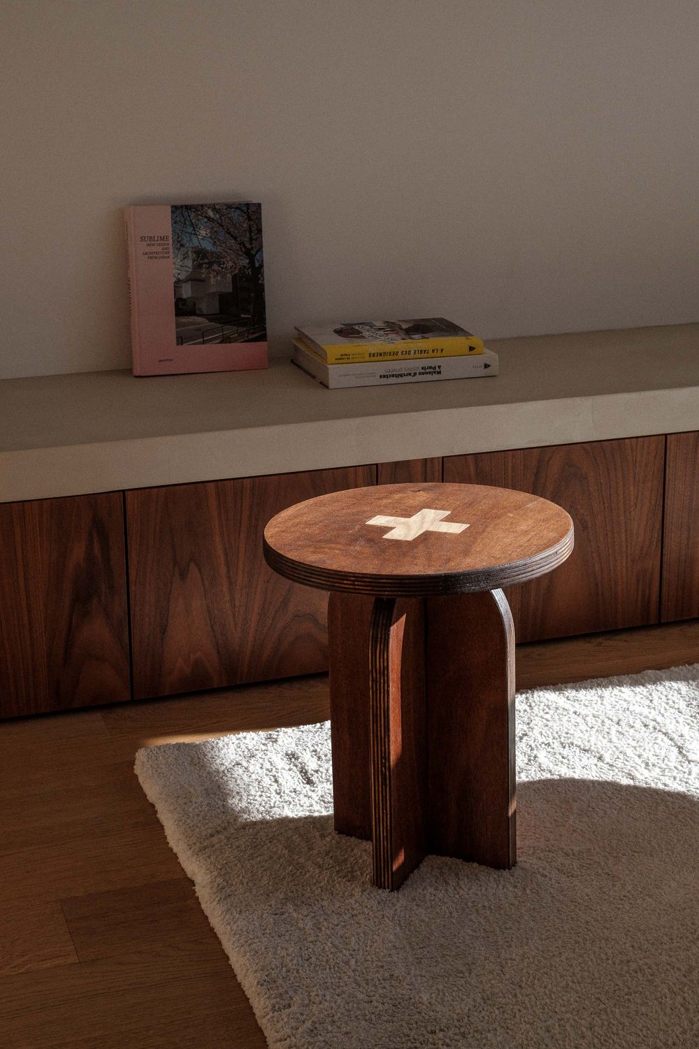 A round wooden side table with a cross detail on top sits on a beige carpet against a wall with wood paneling.