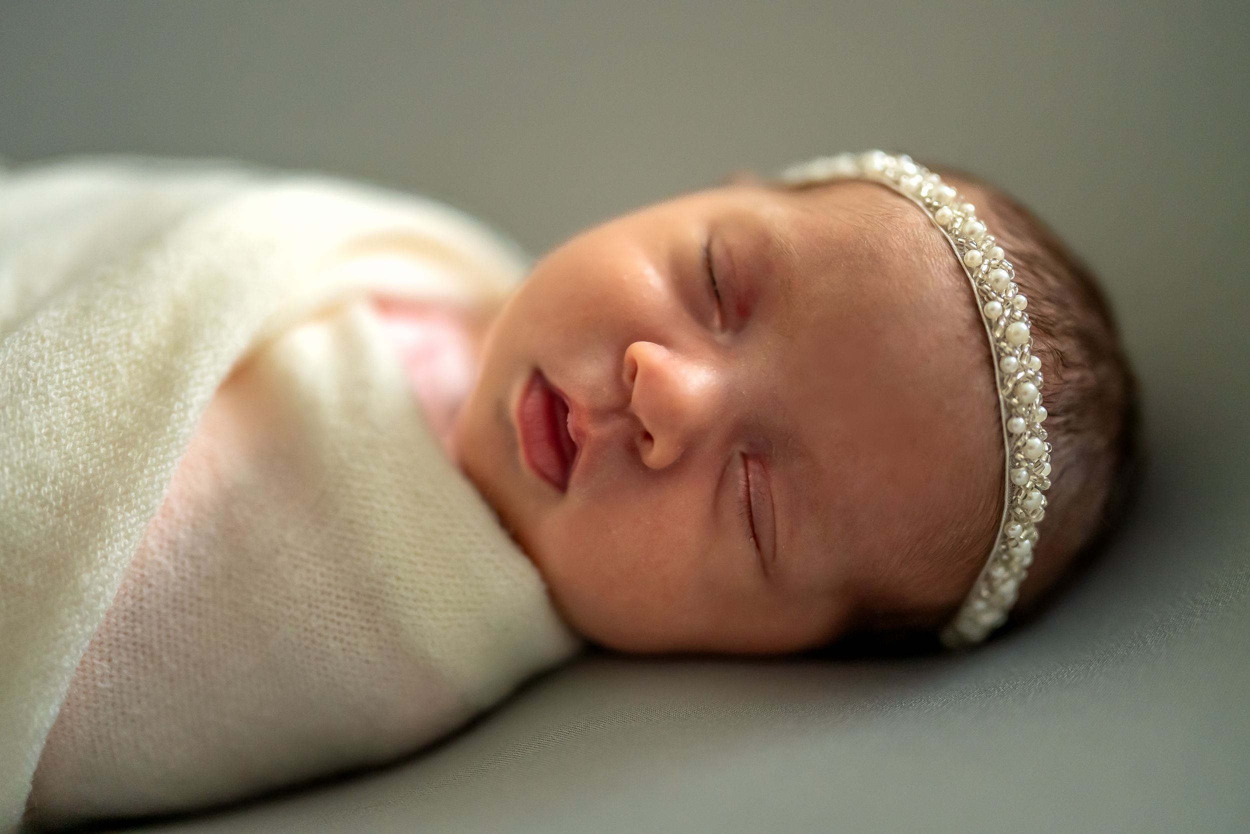 Newborn baby wrapped in soft white blanket with delicate headband resting peacefully.
