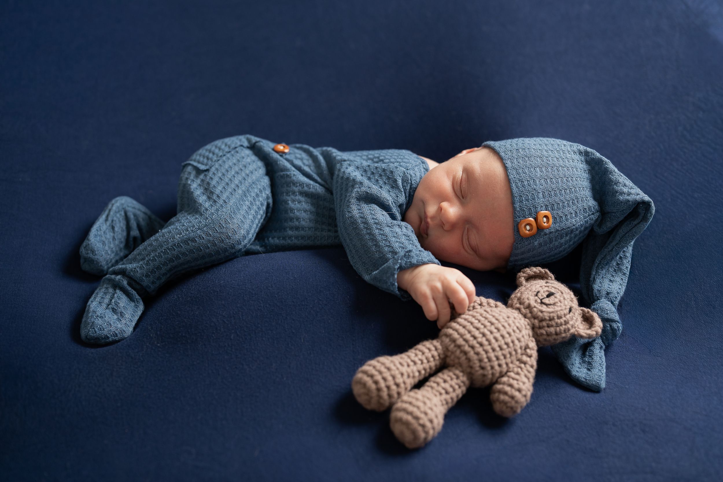 Newborn baby sleeping peacefully in blue knit outfit with matching hat beside a crocheted teddy bear on blue background.