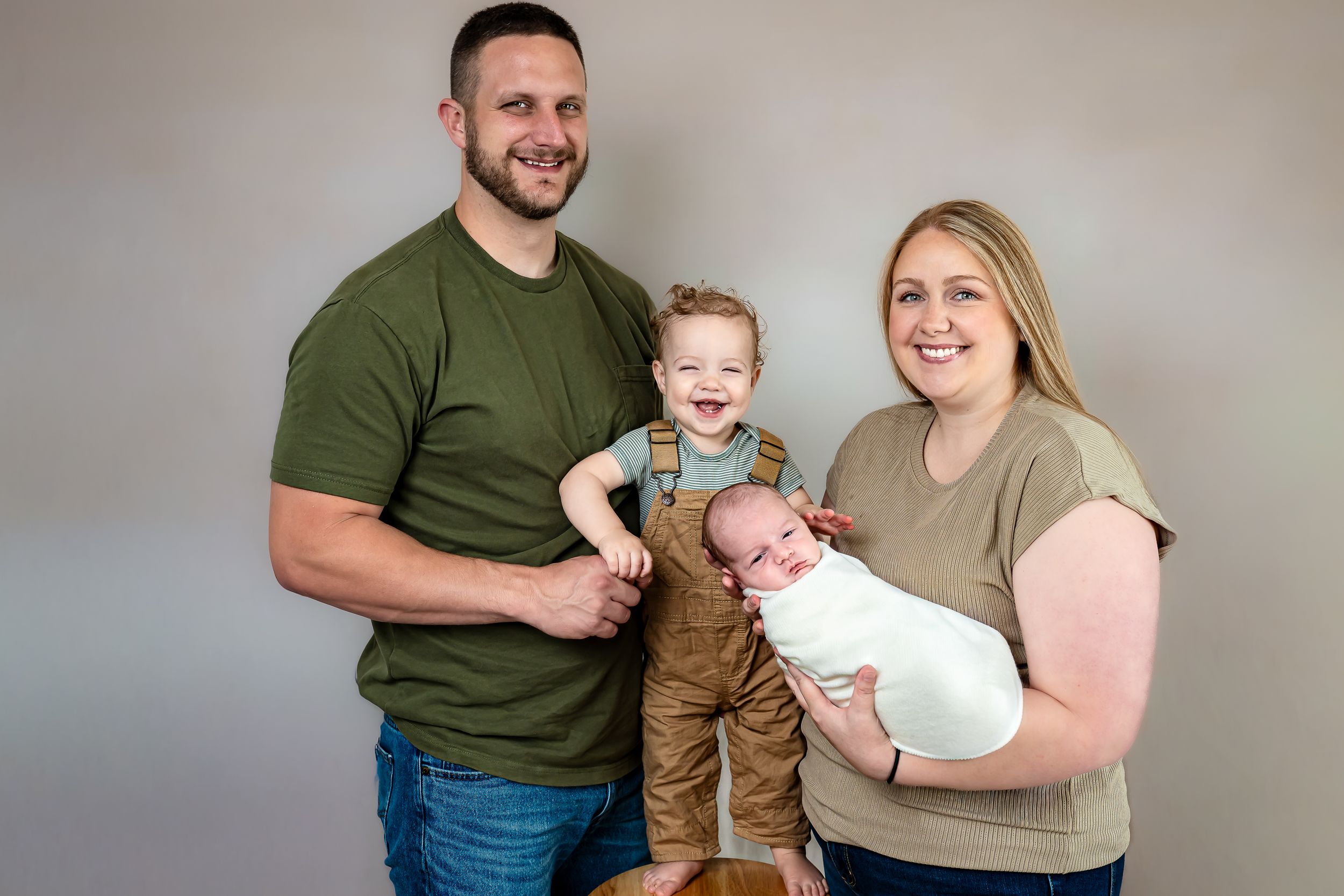Family portrait of parents with toddler and newborn baby posing together against a gray studio background.