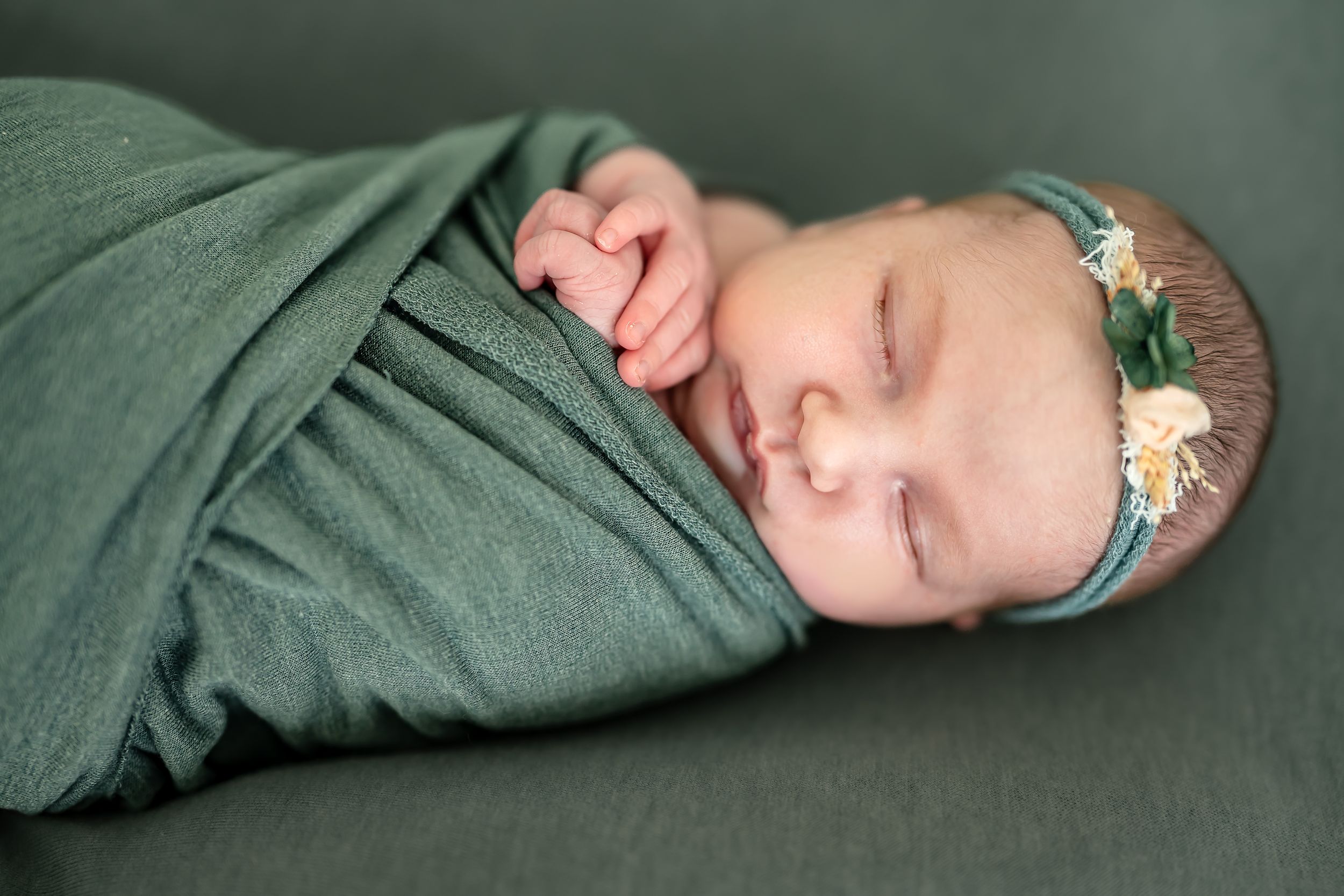 Sleeping newborn baby swaddled in green wrap wearing a floral headband, peaceful infant portrait photography.