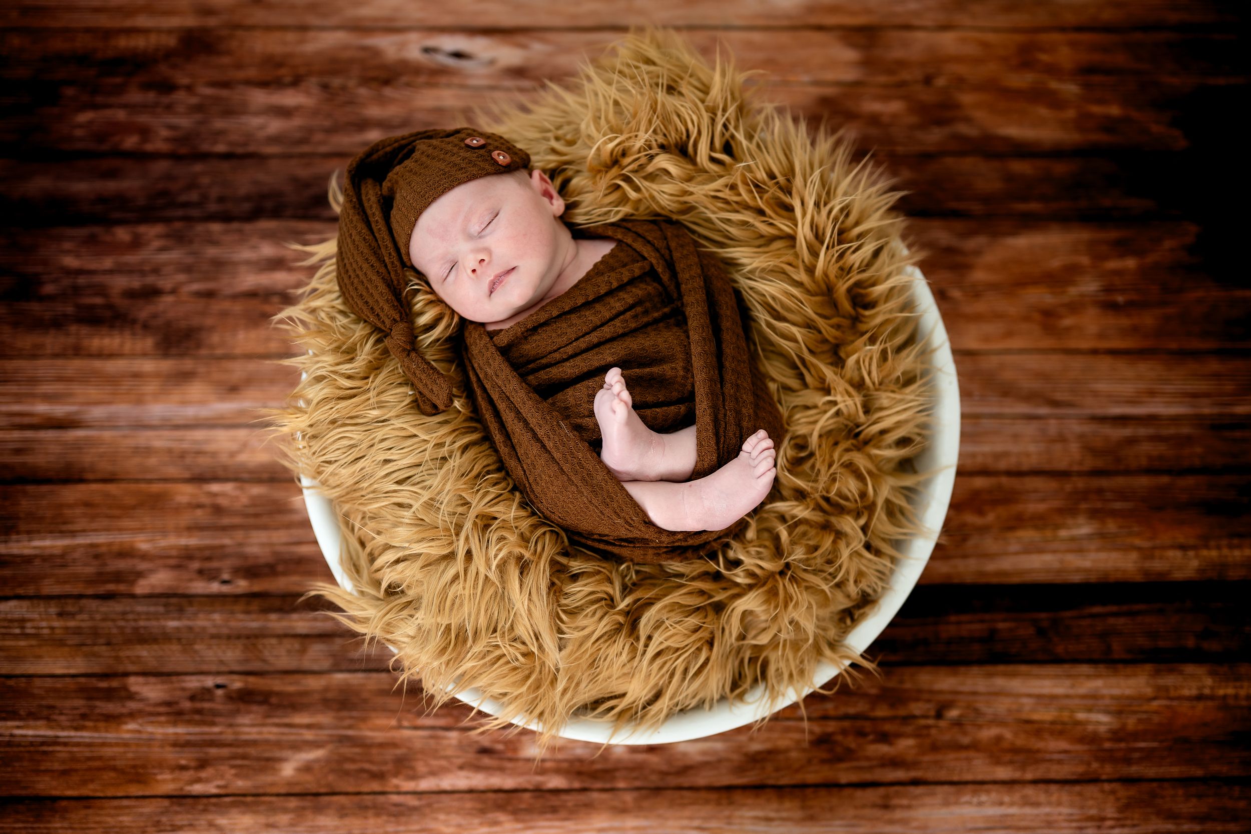 Newborn baby wrapped in brown swaddle sleeping in fluffy tan fur bowl on rustic wood floor.