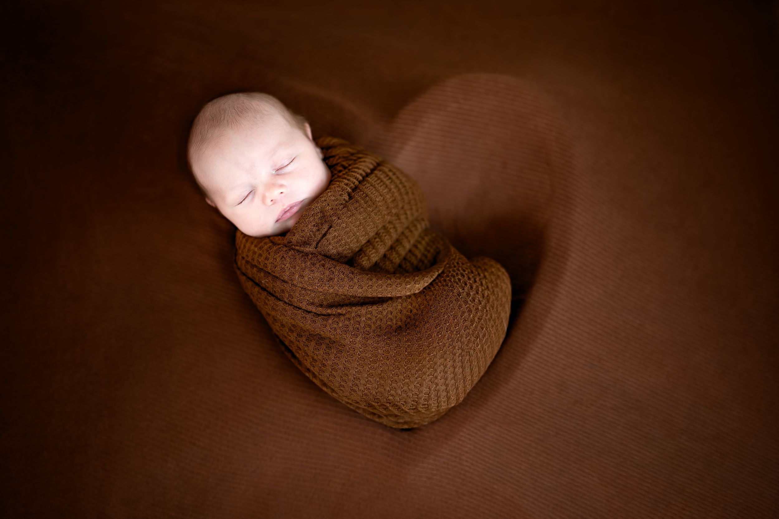 Newborn baby swaddled in brown knit wrap, sleeping peacefully inside a heart-shaped cutout on a brown background.