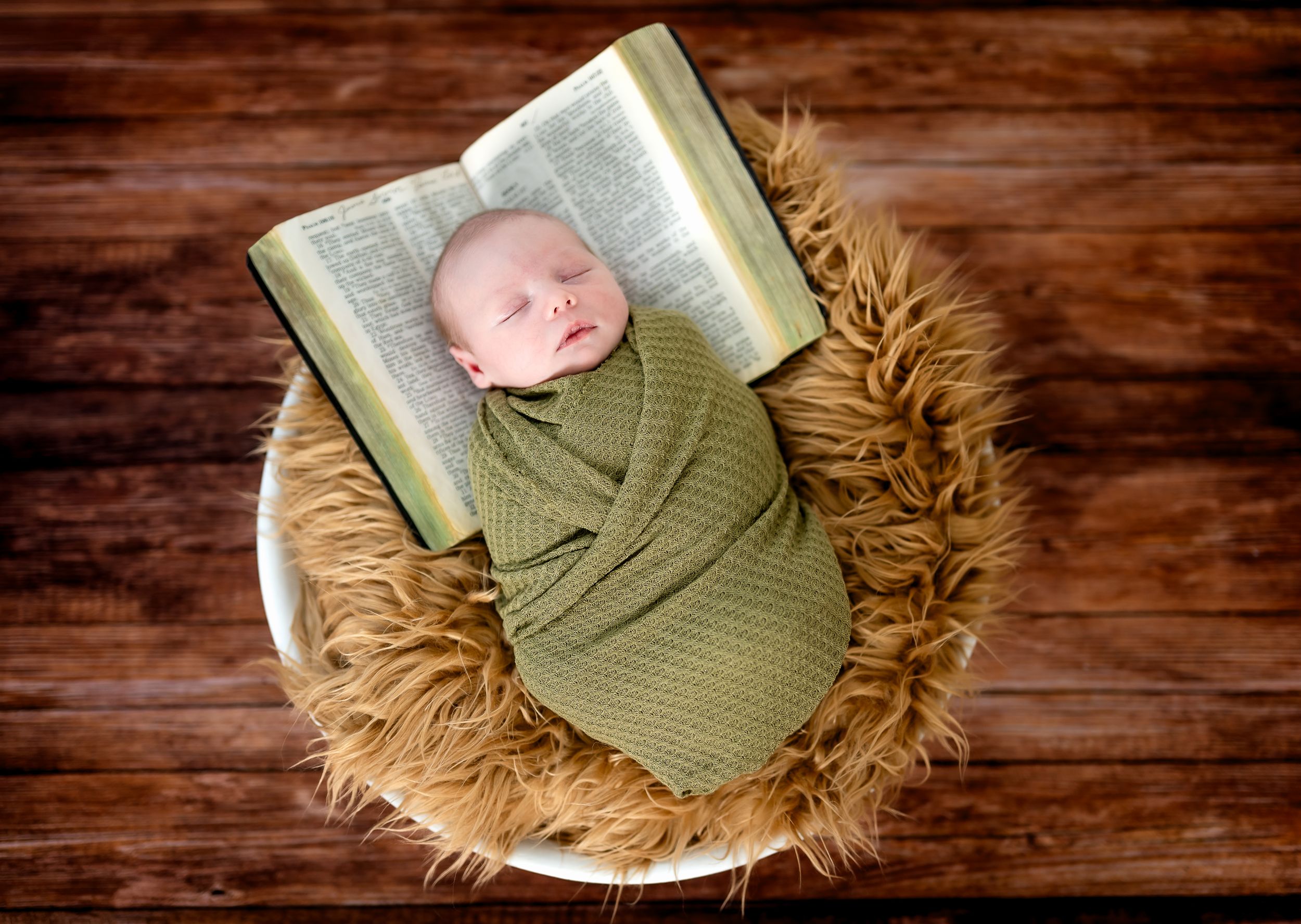Newborn baby swaddled in green wrap sleeping peacefully in a fur bowl with an open Bible.