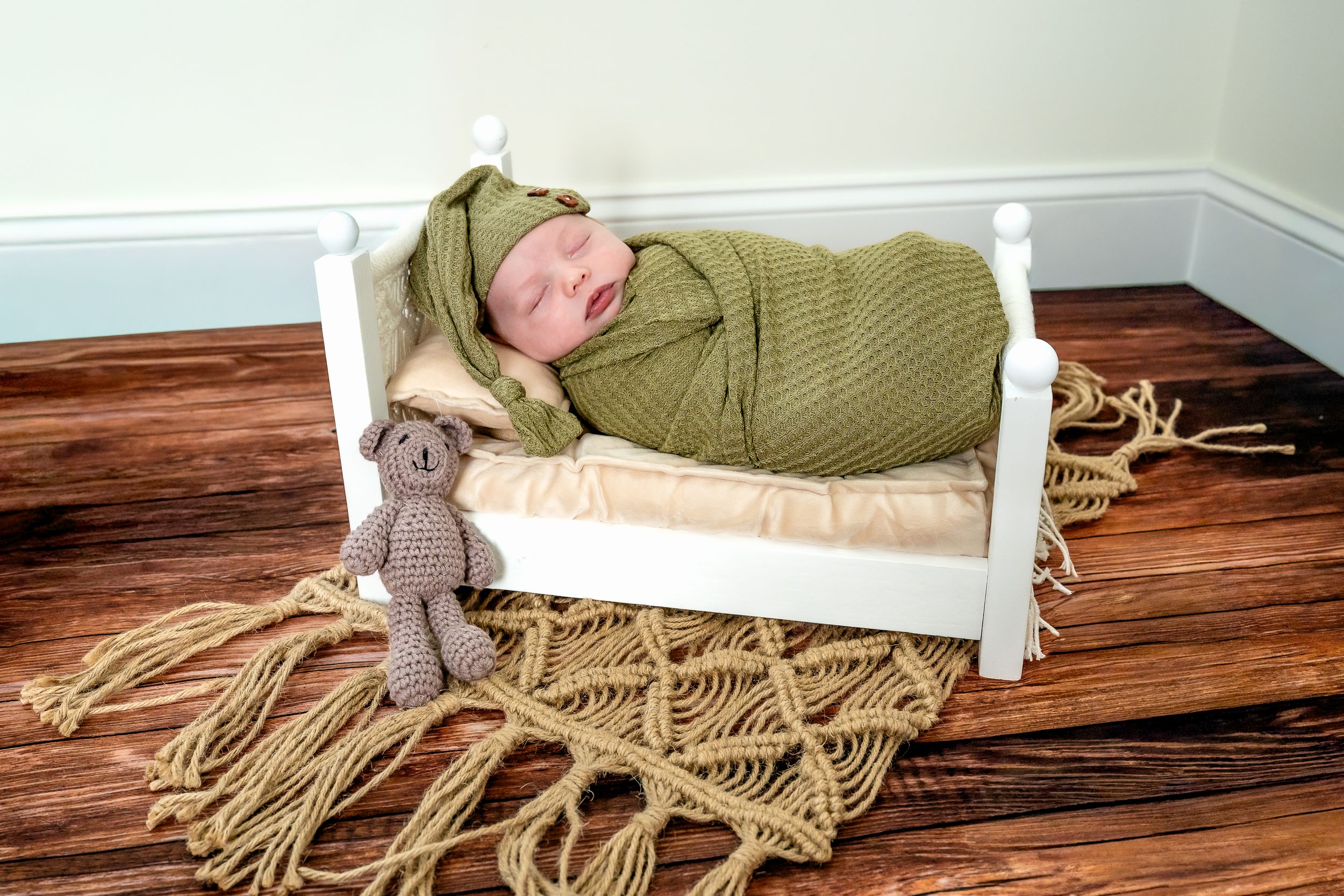 Newborn baby swaddled in green wrap sleeping in a small white wooden bed with a knit teddy bear.