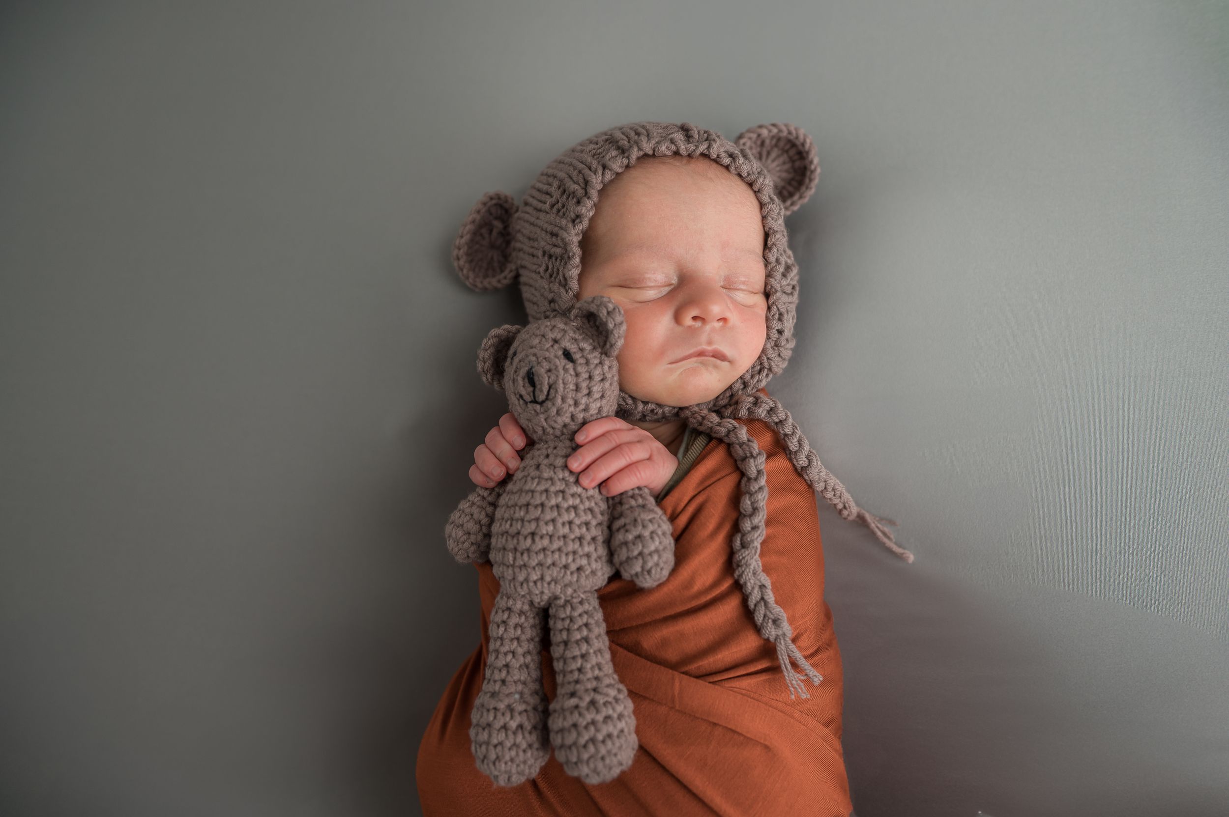A sleeping baby wearing a bear hat and orange outfit while holding a grey knitted stuffed animal.