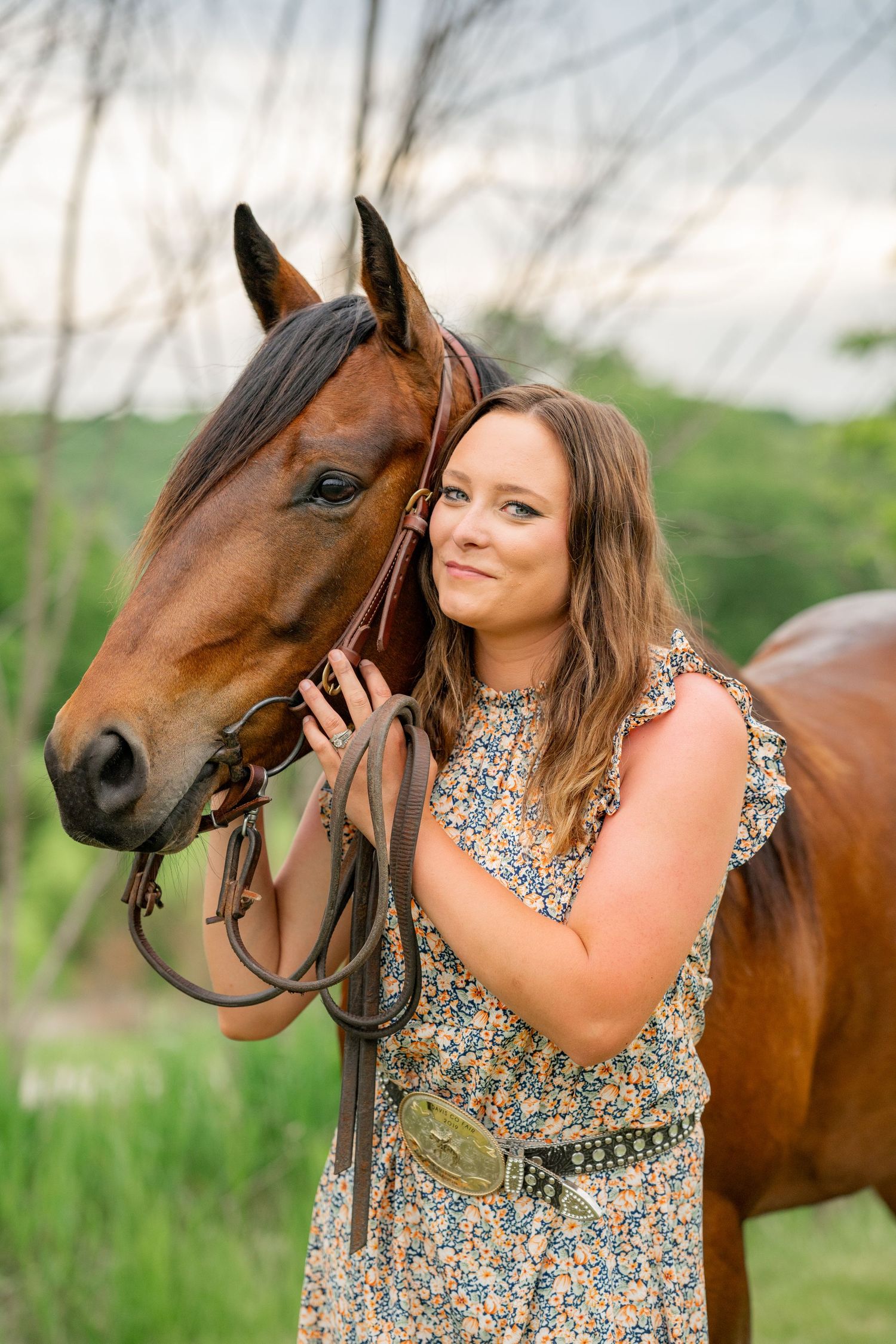 Cheerful woman in floral dress poses affectionately with a beautiful bay horse in a grassy field.
