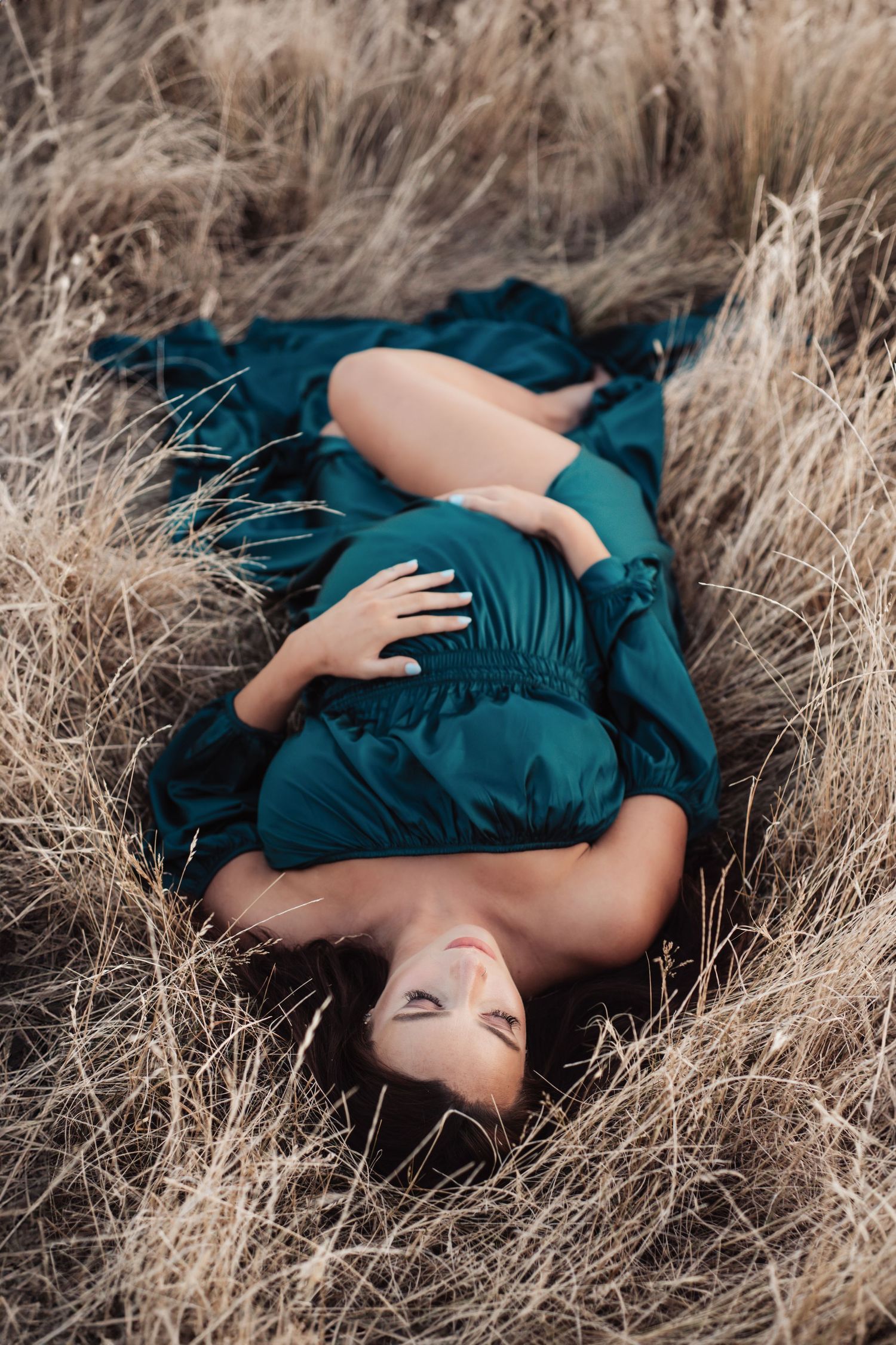A maternity photo of someone laying in tall grass wearing a teal dress.