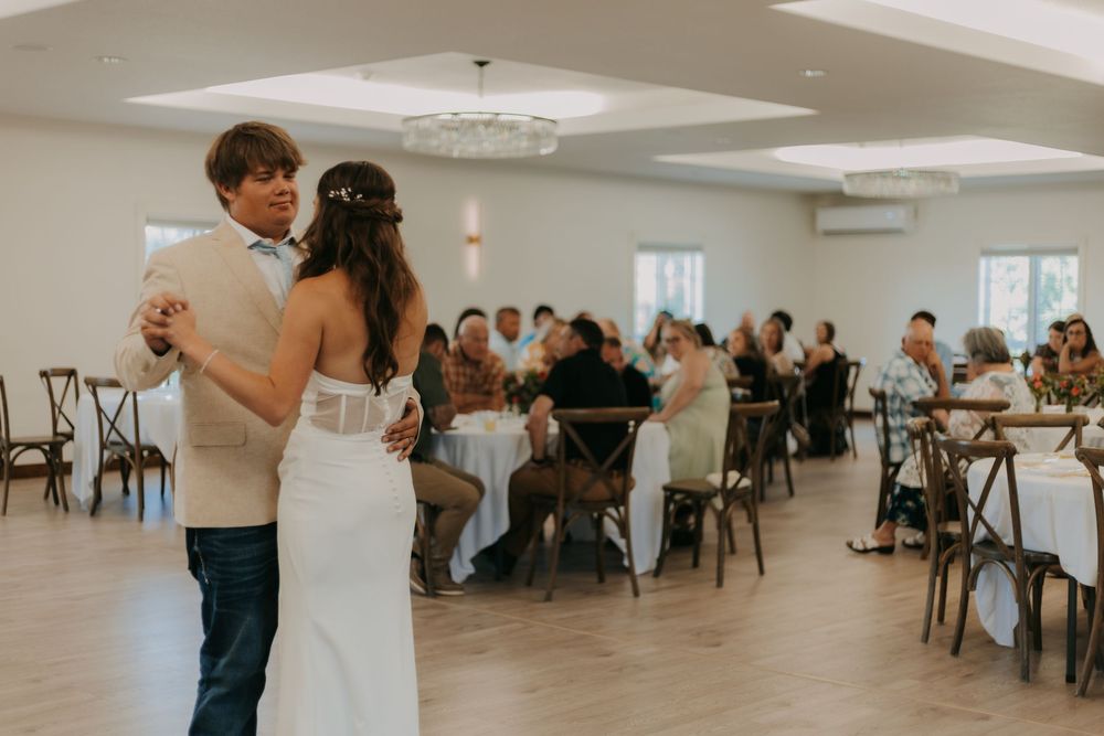 Couple slow dancing in an elegant white reception venue with guests seated at tables in the background.