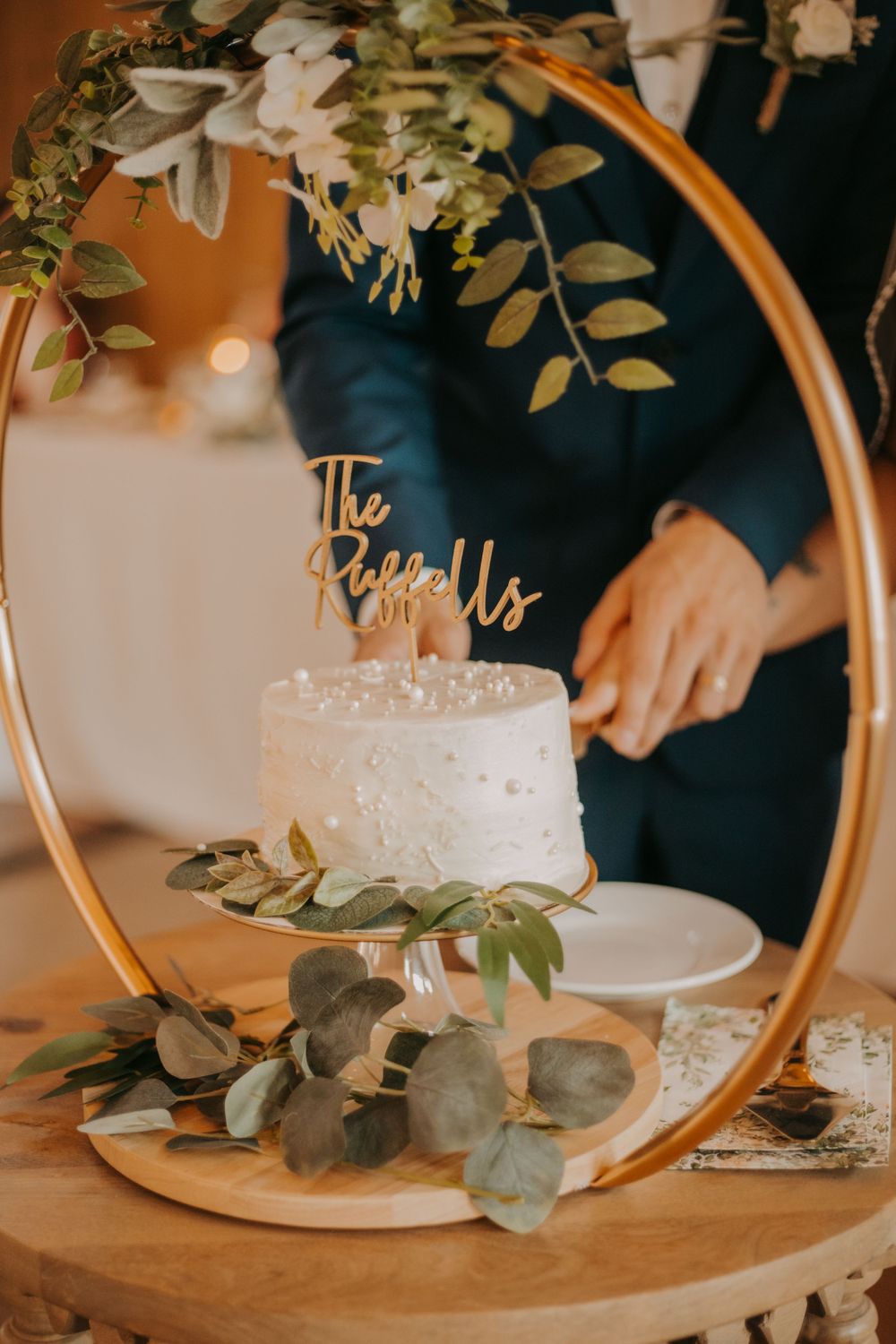 White wedding cake displayed on gold circular stand with eucalyptus and floral decorations.