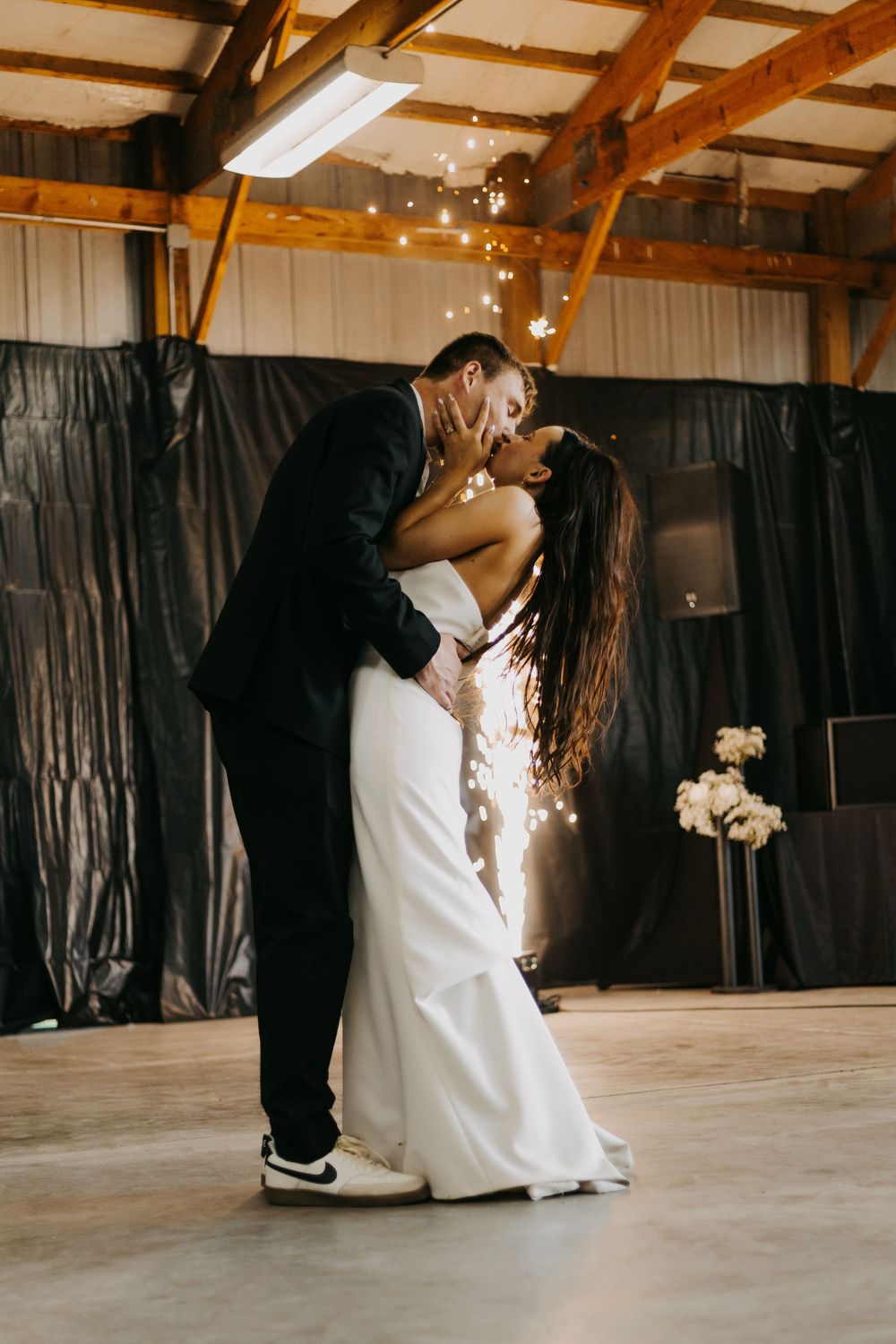 Romantic first dance between newlyweds in a rustic barn venue with exposed wooden beams and string lights overhead.