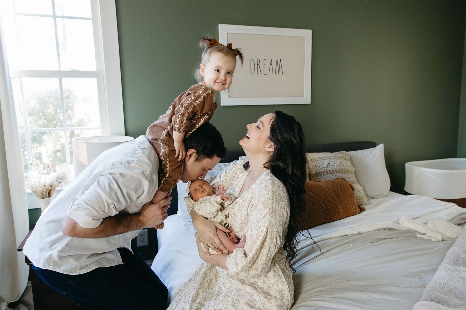 A family plays and laughs together while spending quality time on a bed in a room with green walls and natural light.