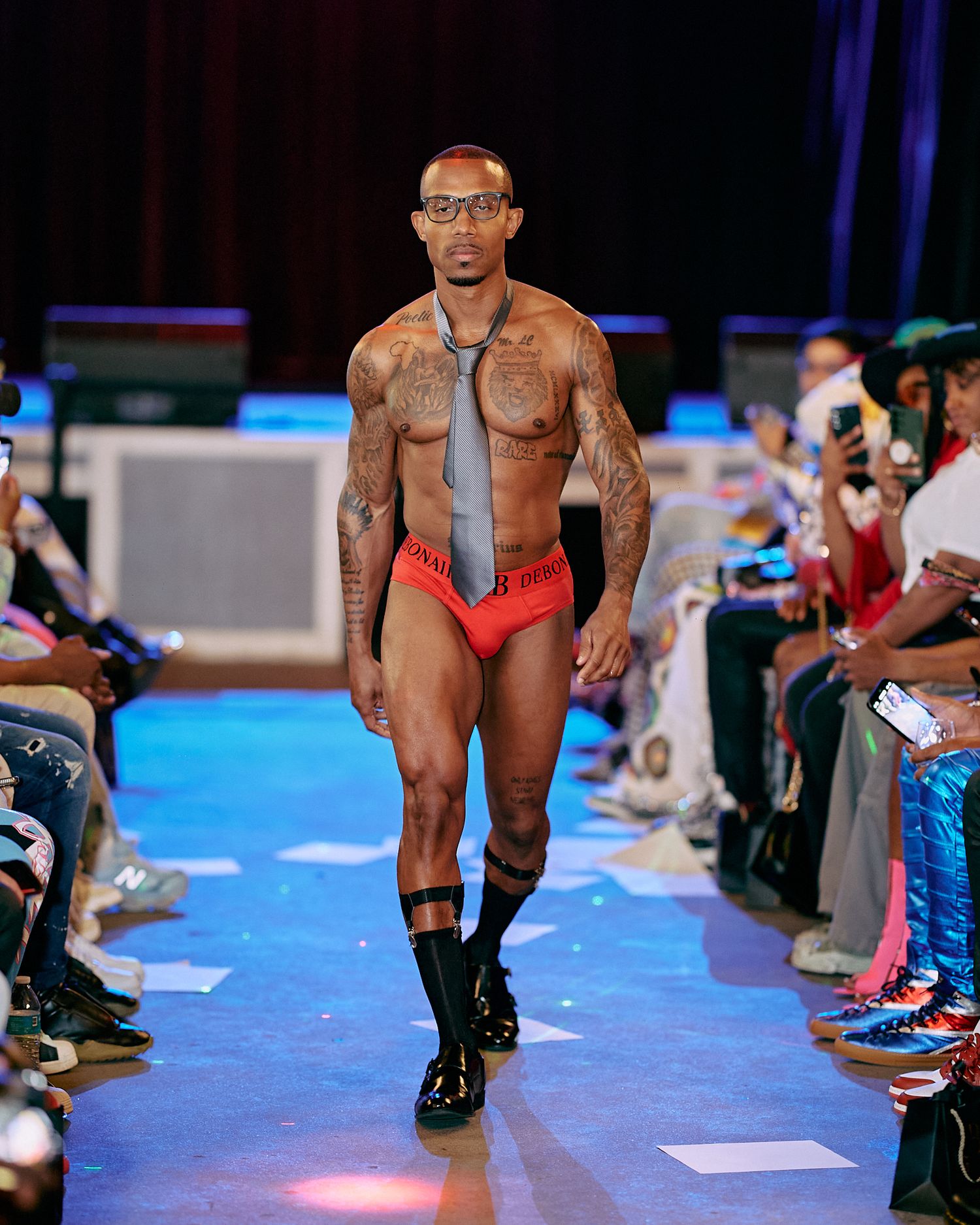 Model walks runway in brown leather jacket and red briefs during fashion show with blue lighting effects.