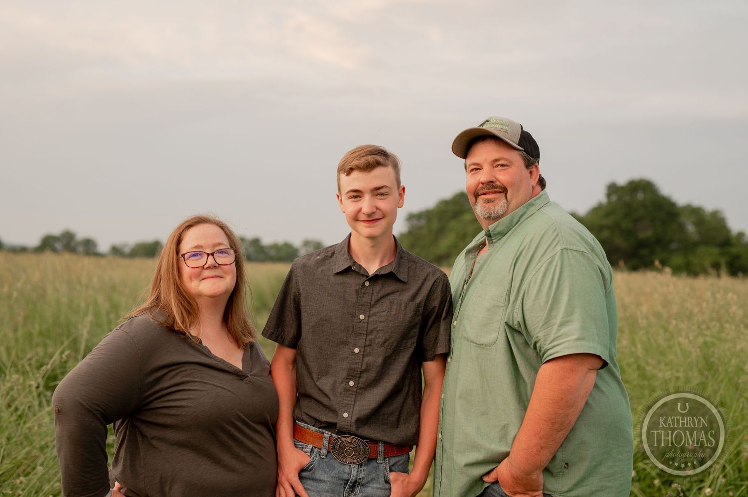 Family of three poses together in a grassy field at sunset with a rustic backdrop.