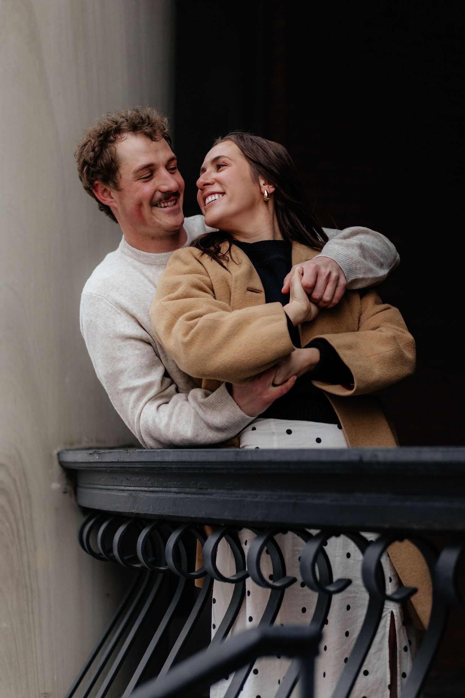 A romantic couple embraces and laughs together while standing on a dark metal balcony.
