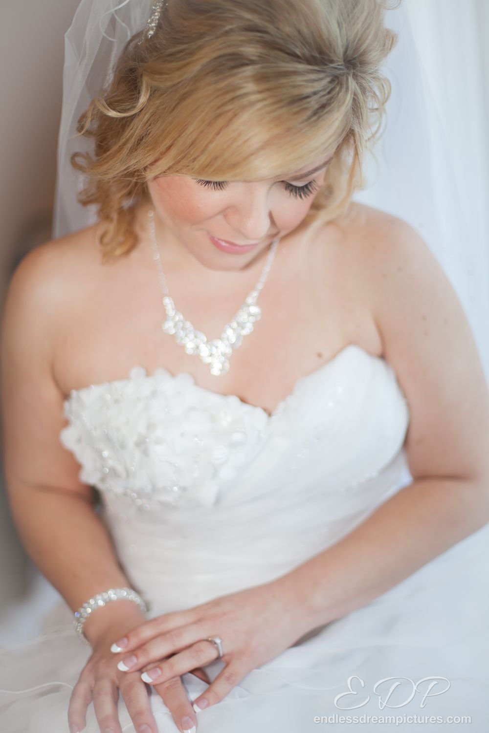 Bride in white wedding dress and jewelry looking down with a gentle smile.