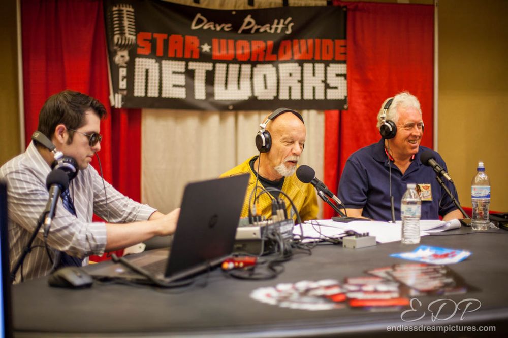 Three radio hosts wearing headphones broadcasting from studio with red curtain backdrop.