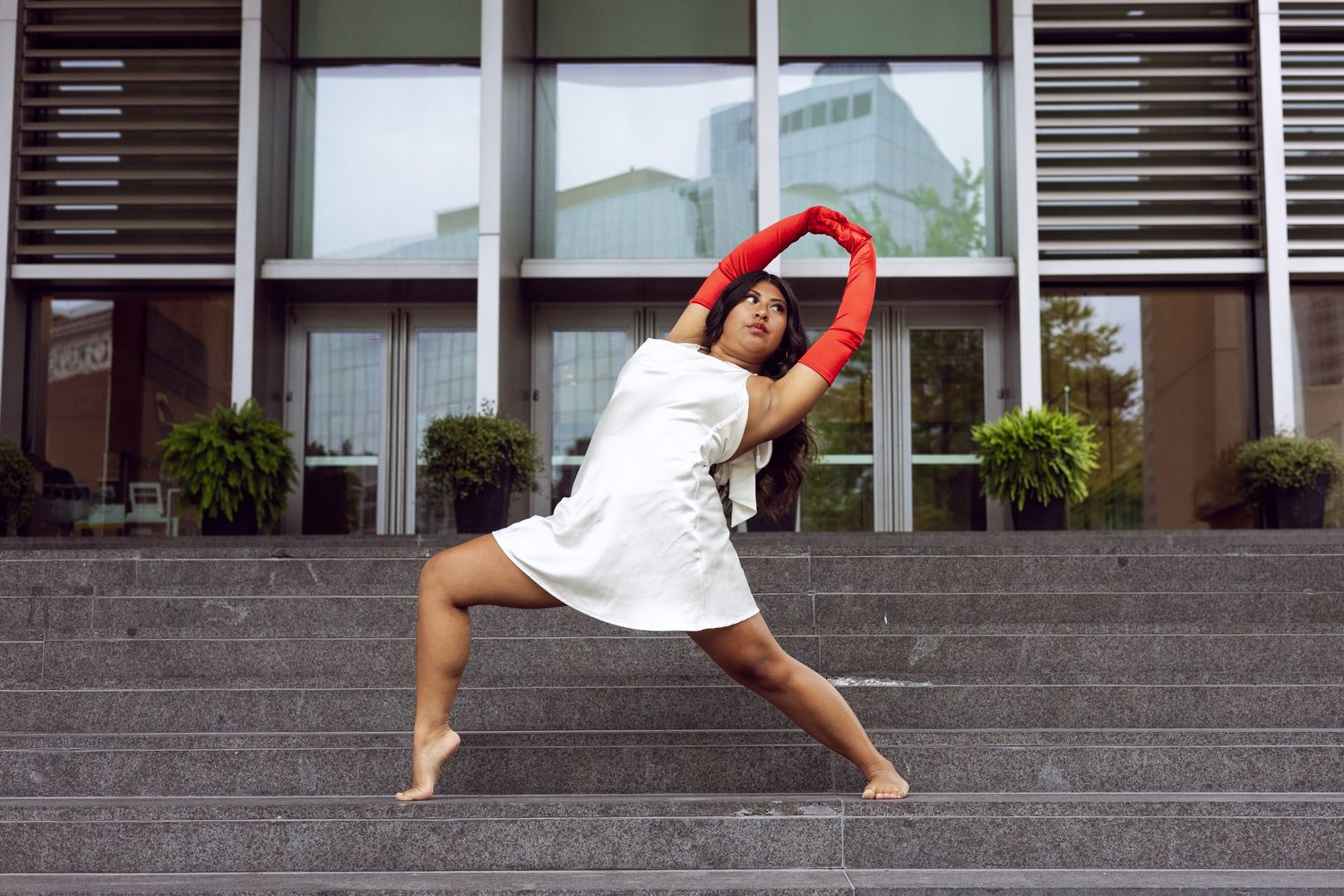 A dancer in a white dress performs a graceful pose on concrete steps outside a modern building. Grand Rapids.