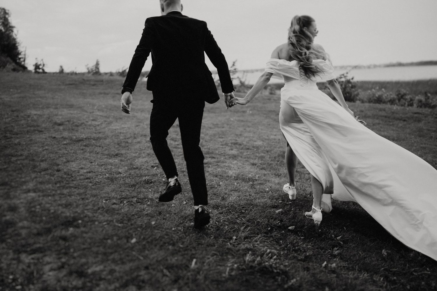 Black and white photo of newlyweds running playfully across a field with flowing dress.